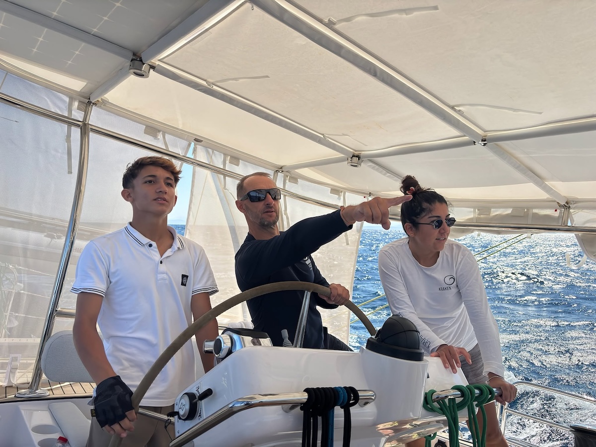 Zwei Crewmitglieder sch&uuml;tzen sich mit lang&auml;rmliger Funktionskleidung. Alle geniessen das Segeln in der sonnigen &Auml;g&auml;is im Schatten der Pergola